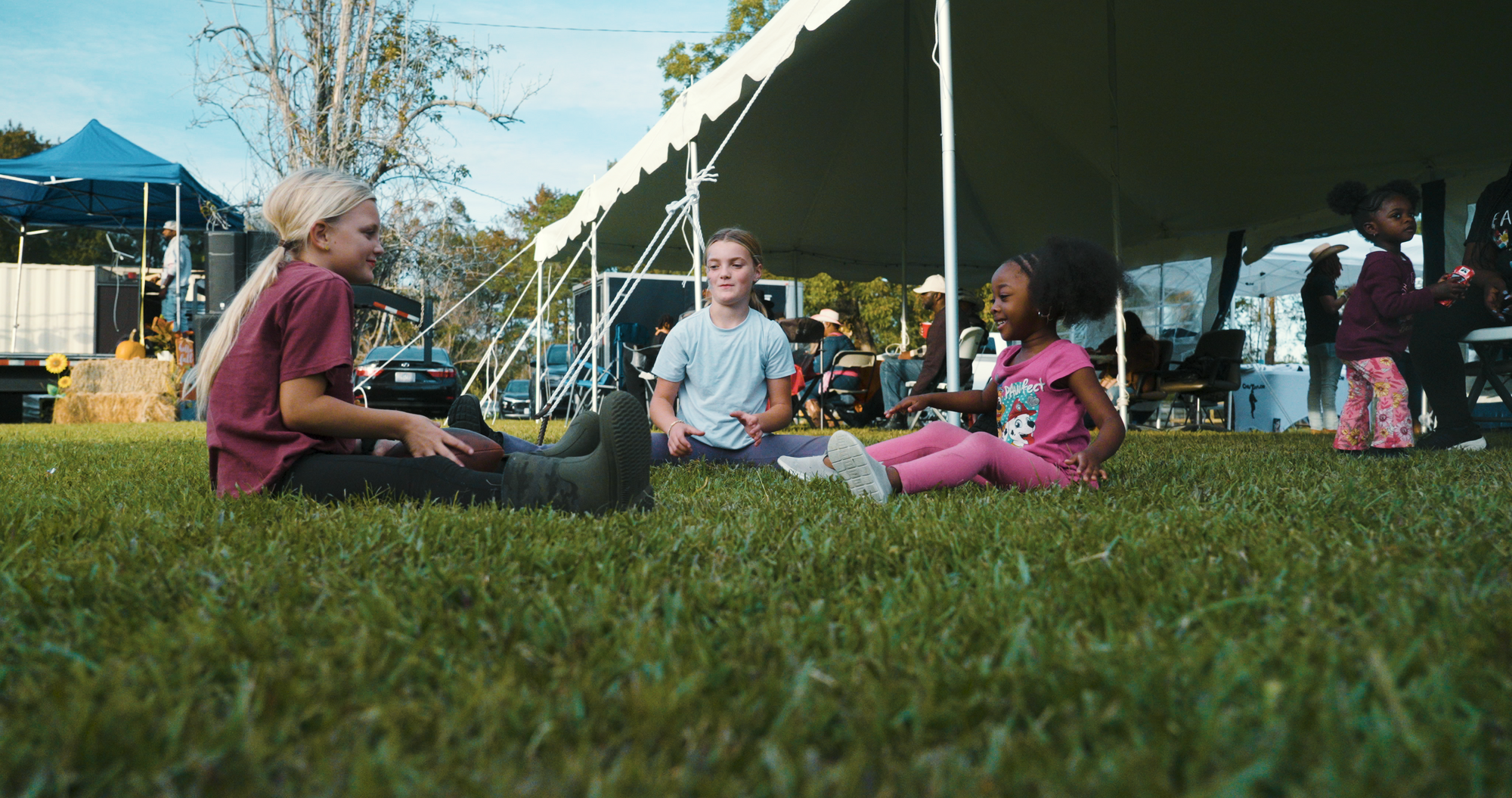 People sitting on grass under a large white tent with event activities in the background